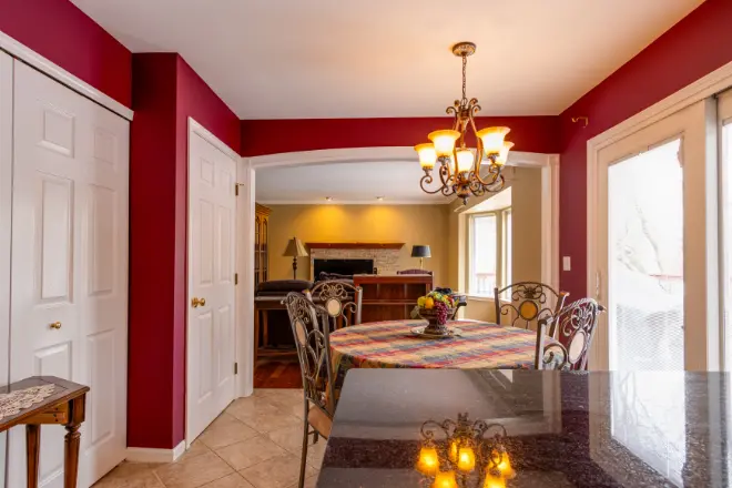 A beautifully designed dining area in Southeast Michigan, expertly painted by Tribble Painting, features a round table with chairs beneath a sparkling chandelier. Rich red walls, white closet doors, and elegant tile flooring create a welcoming atmosphere. Ample natural light pours in through large glass doors, showcasing the high-quality interior painting—perfect for homes in Ann Arbor, Ypsilanti, or Novi. by Tribble Painting